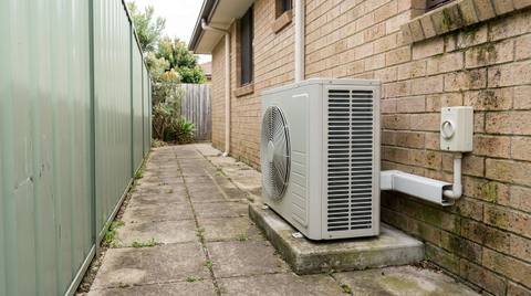An air conditioning outdoor unit installed on a concrete slab in the side passage of an Australian brick veneer home, beside a Colorbond fence